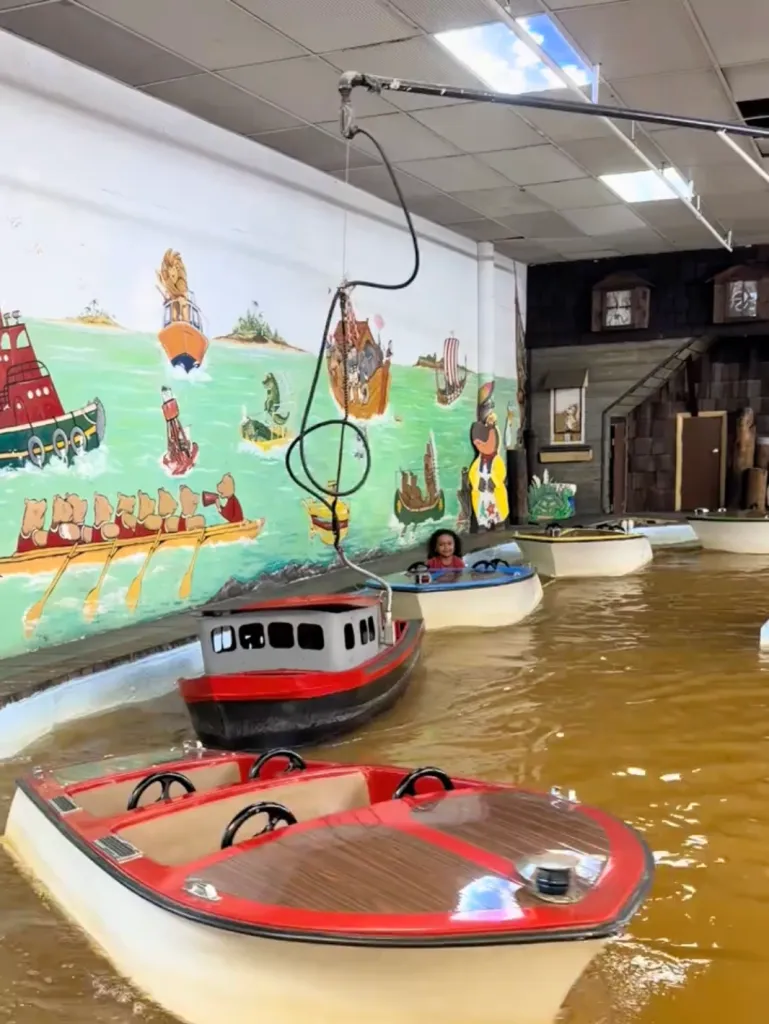 A child riding a small boat in a pool of murky brown water at a theme park, next to a mural of cartoon boats.