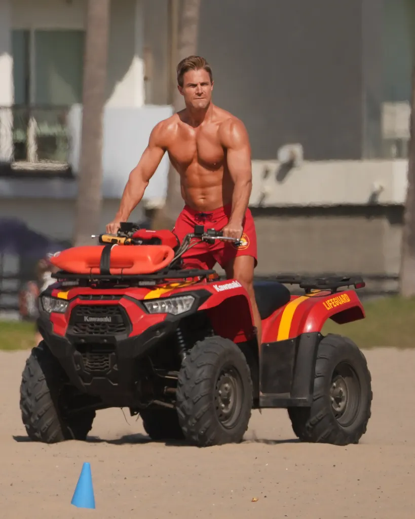 Stephen Amell, shirtless and wearing red shorts, drives a red and yellow lifeguard ATV on the beach.