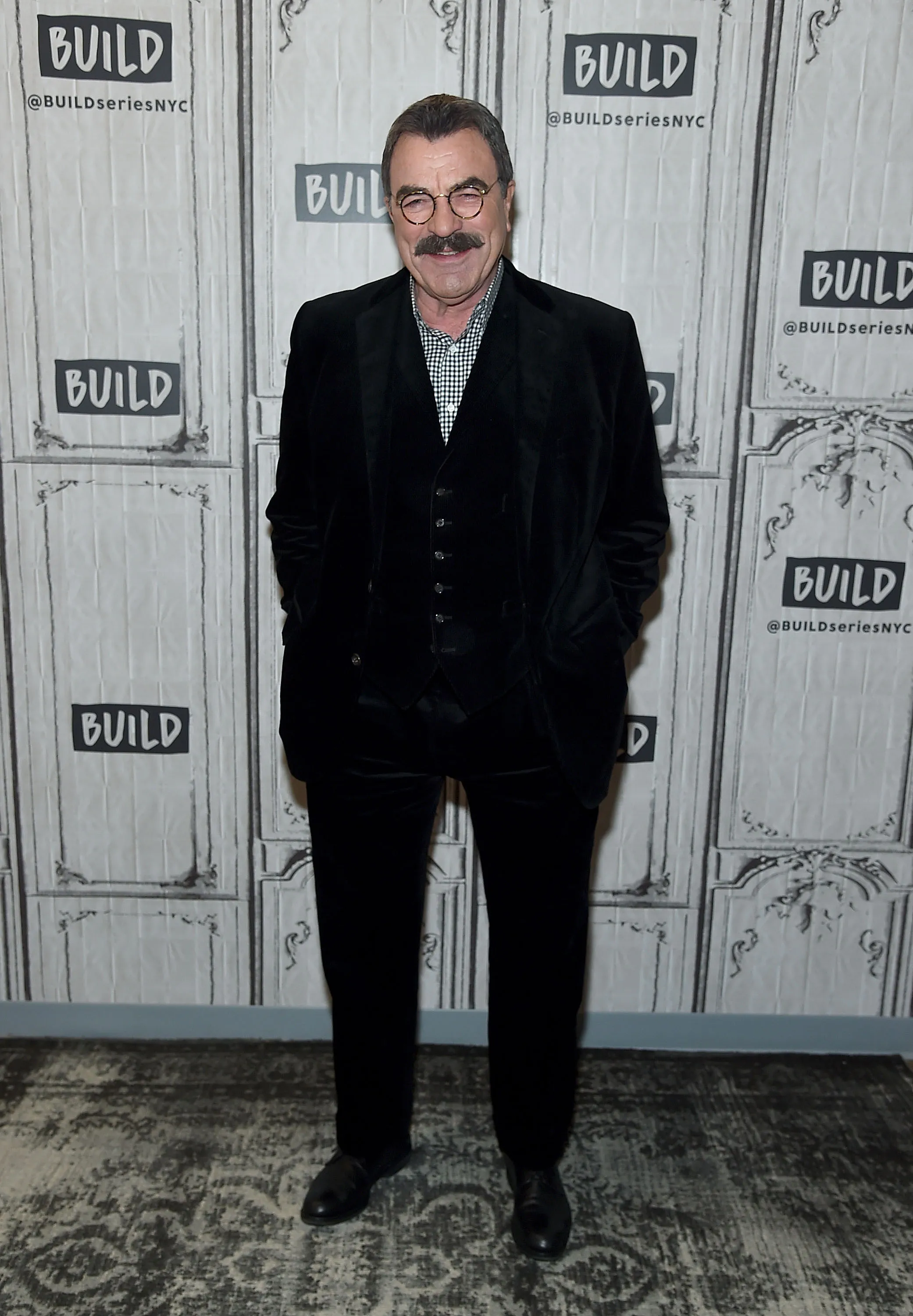 Tom Selleck smiling in a black velvet suit, a white and black checkered shirt, and round glasses, standing against a backdrop of white panels with the word