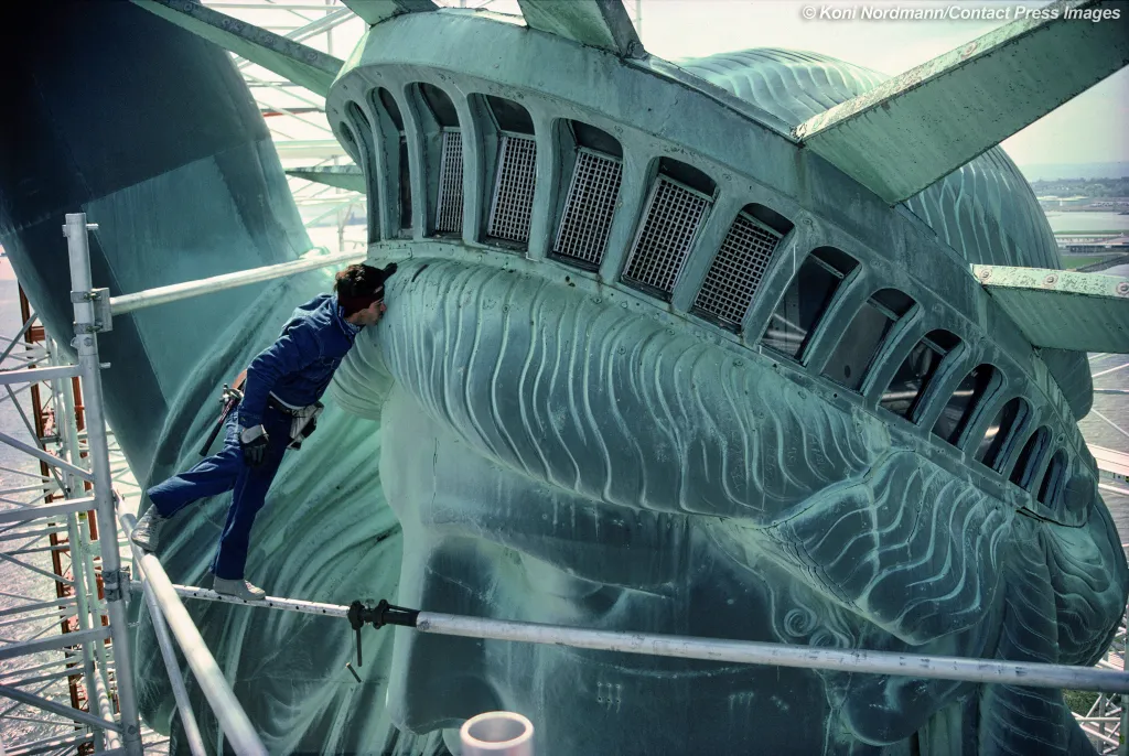A man in work clothes leans in to kiss the Statue of Liberty's cheek, which is weathered and green.