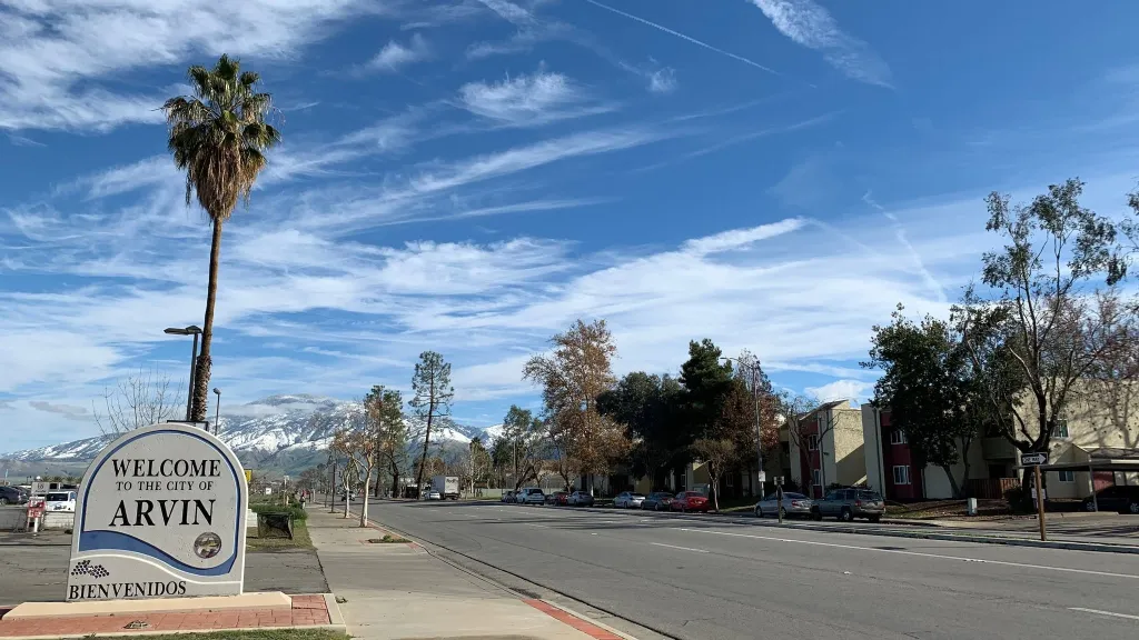Sign welcoming visitors to Arvin, with a palm tree, street, buildings, and snow-capped mountains in the background.