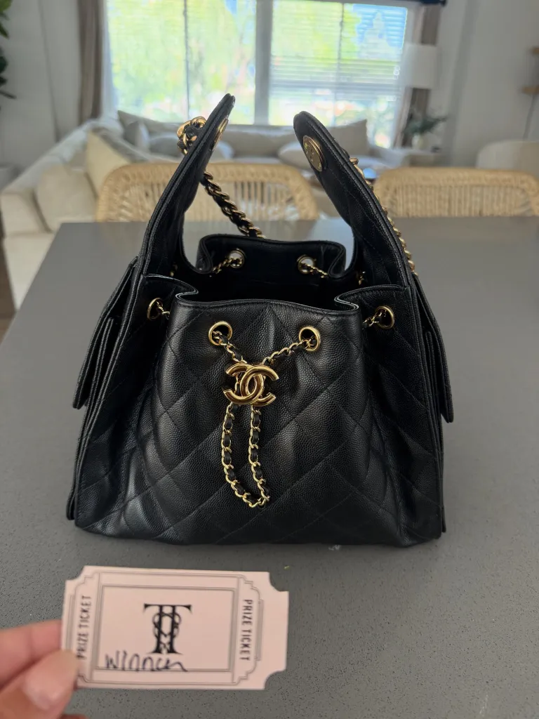 A black quilted Chanel handbag with gold chains sits on a gray counter, next to a hand holding a pink raffle ticket with 