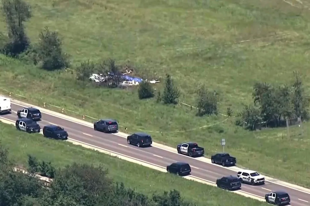 Aerial view of a plane crash site near Butler Memorial Airport, with wreckage covered by a blue tarp and police vehicles on a nearby road.