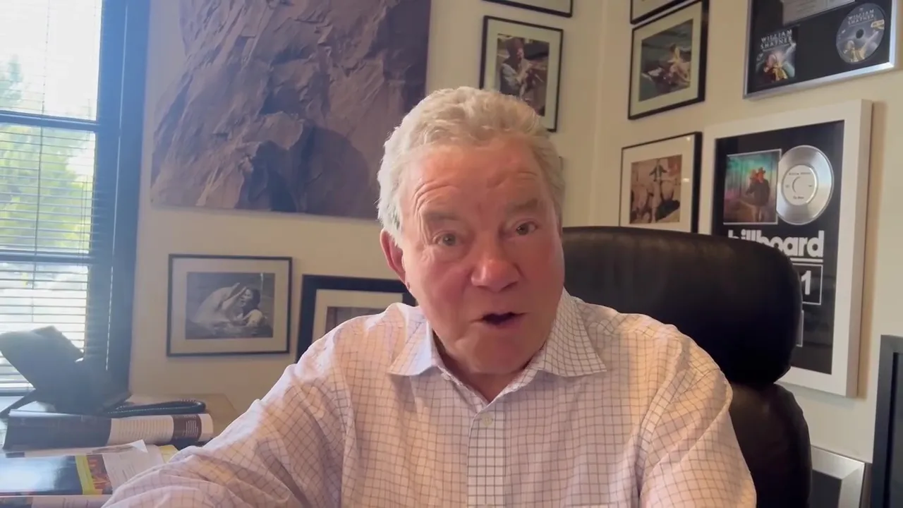 William Shatner in a plaid shirt with an open mouth, sitting in an office with framed awards and artwork on the wall.