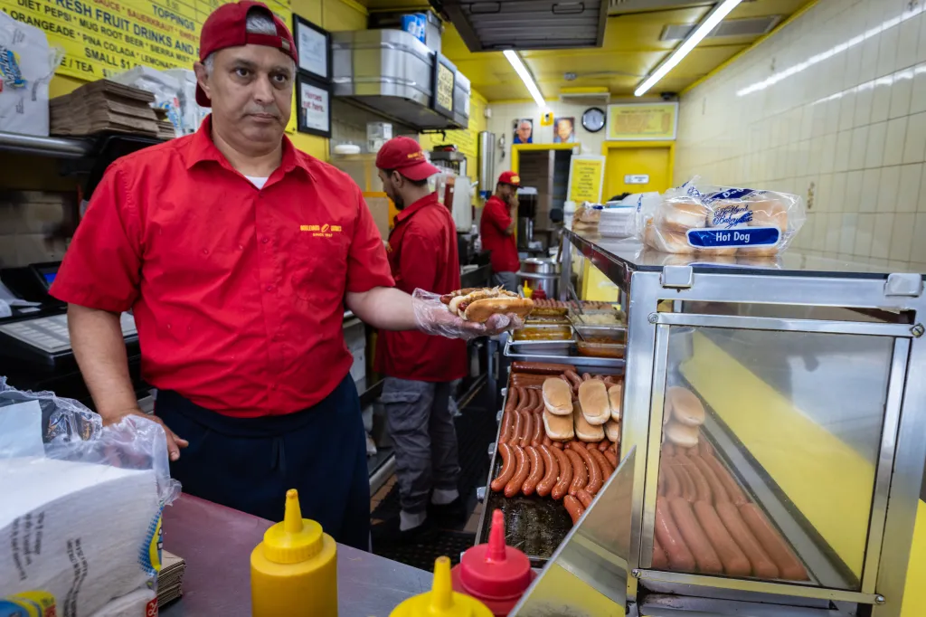 A worker in a red shirt holding two hot dogs in a bun, with a hot dog stand visible behind him.