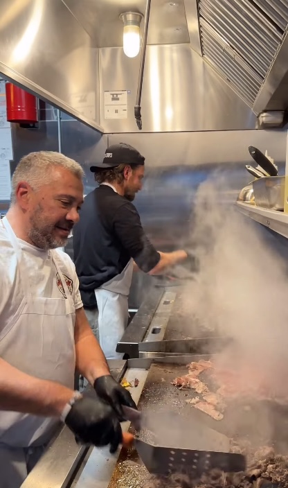 Two men cooking meat on griddles in a commercial kitchen with steam rising.