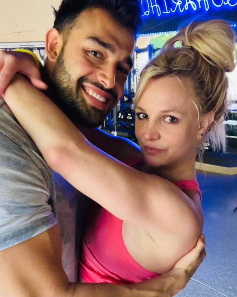 Sam Asghari, in a grey T-shirt, miling at the camera while embracing Britney Spears, in a red tank top, who is looking over his arm at the camera.