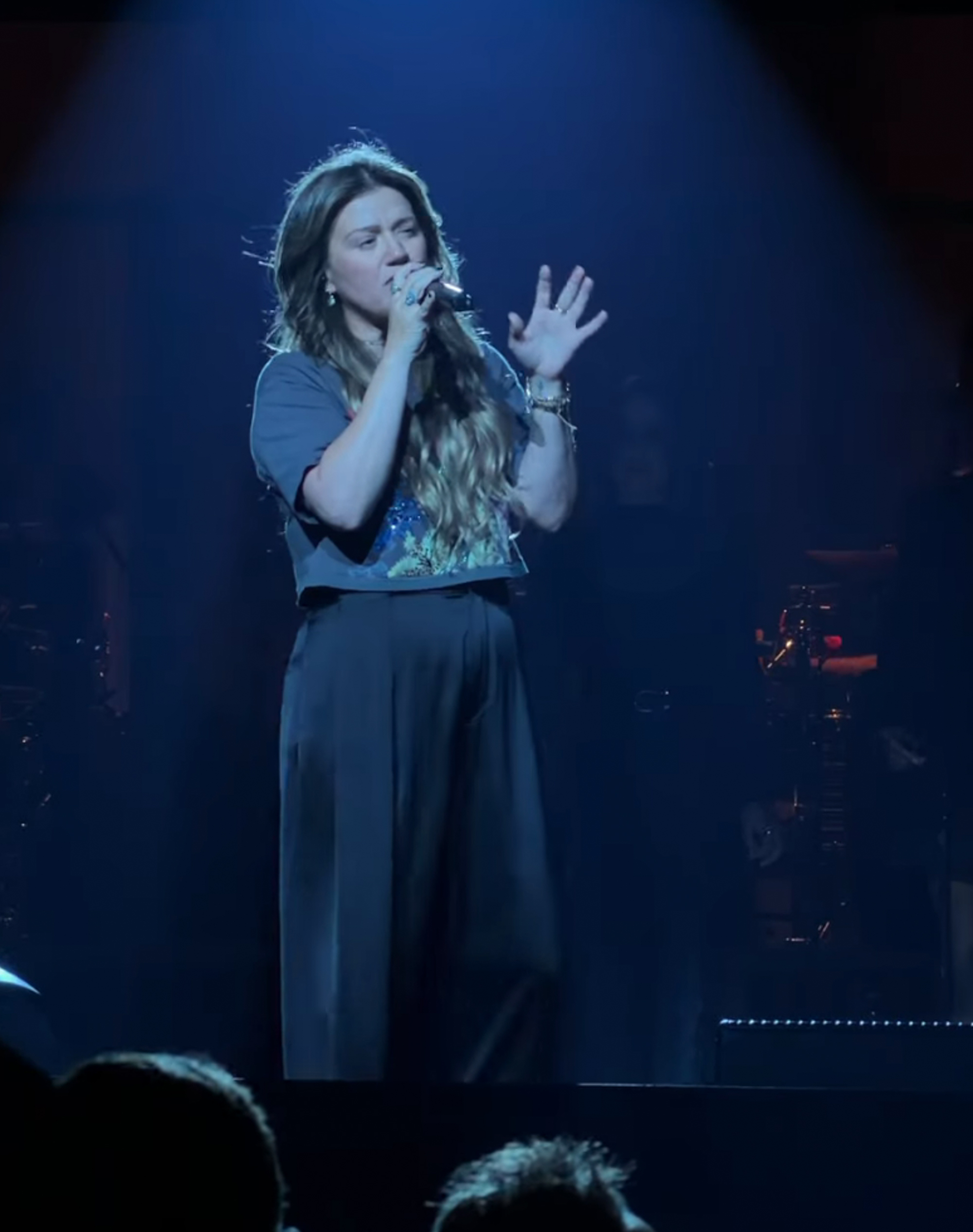Kelly Clarkson singing into a microphone on stage under a blue spotlight.