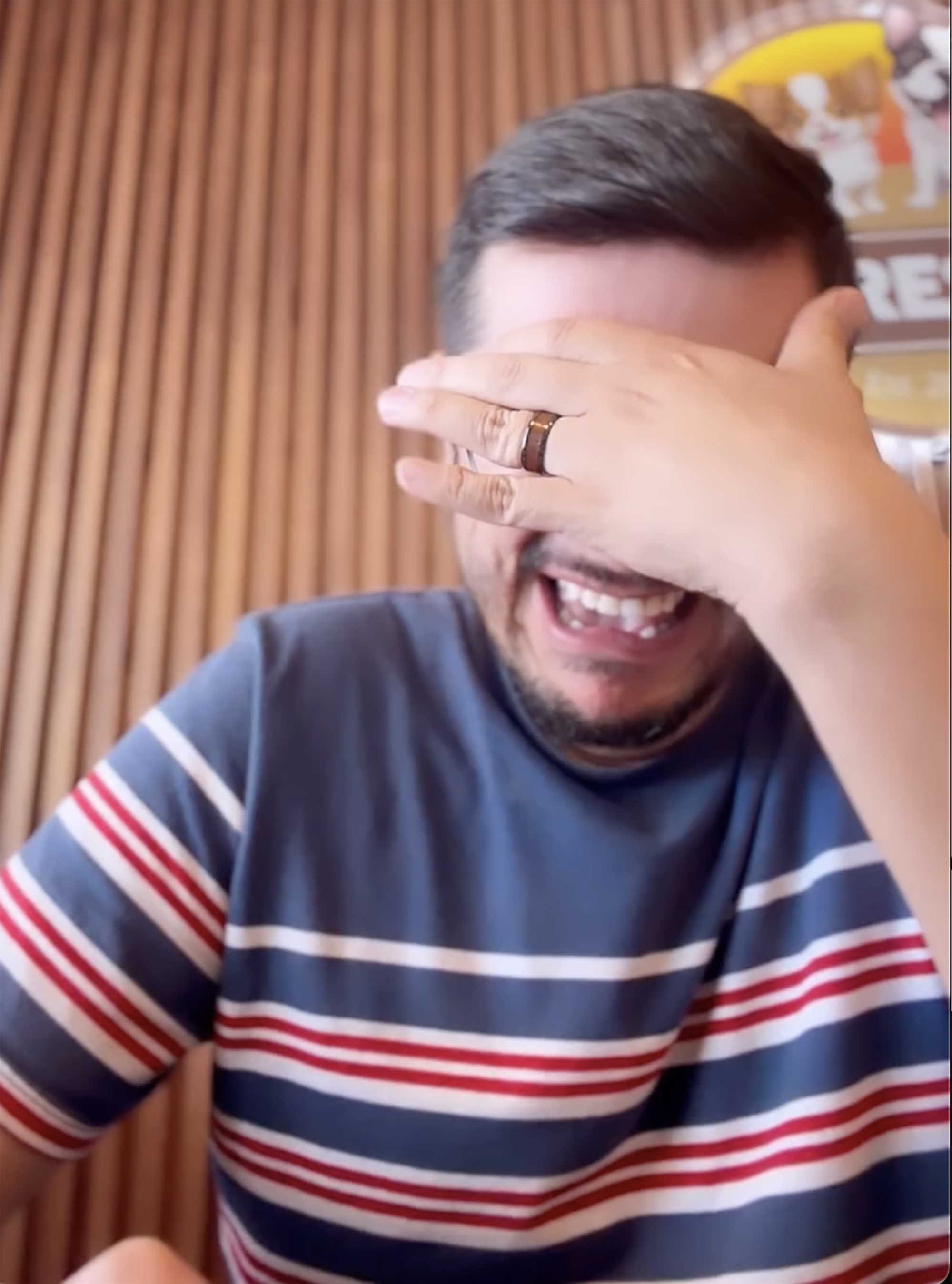 A man in a striped shirt laughs with his hand over his eyes.