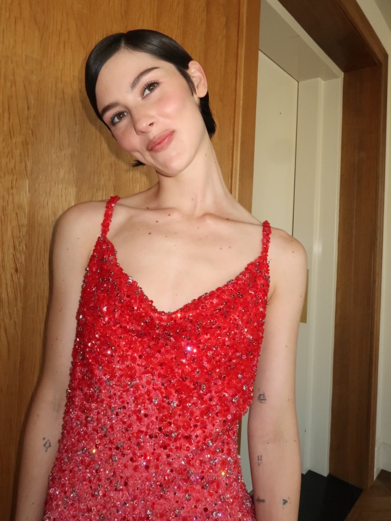 Gracie Abrams in a red sequined dress, smiling playfully.