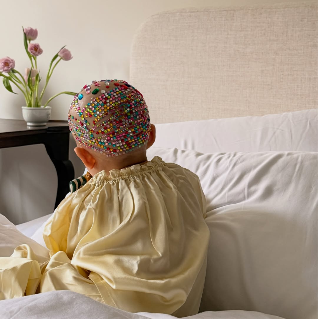 Child in a yellow smock with a jeweled head covering, sitting up in bed.
