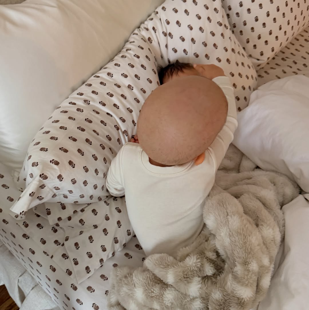 Overhead shot of two babies sleeping in a bed, one with a bald head in a white onesie, and the other with dark hair next to a pillow.