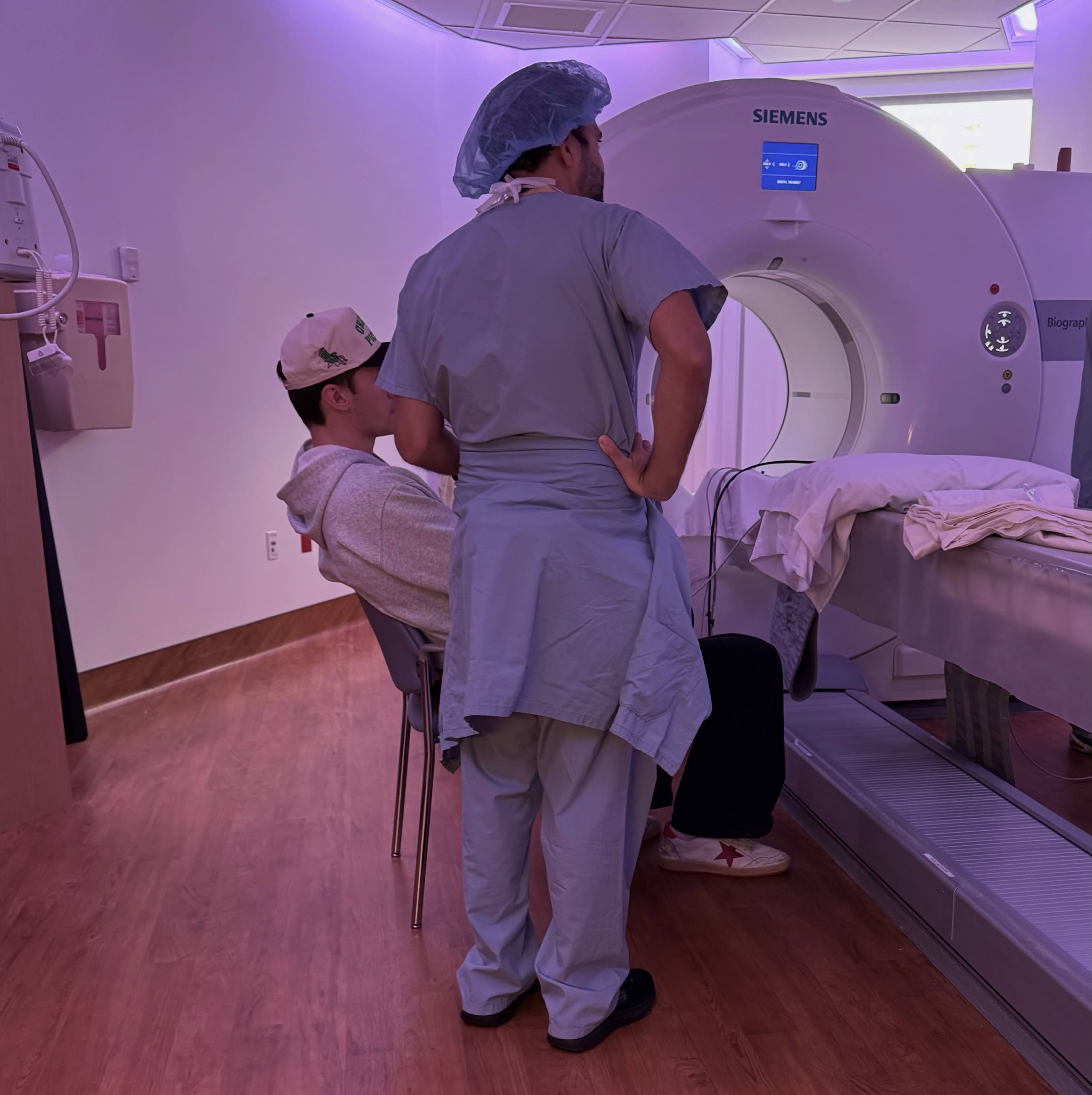 A person in scrubs and a surgical cap standing next to a patient sitting by a Siemens medical imaging machine.