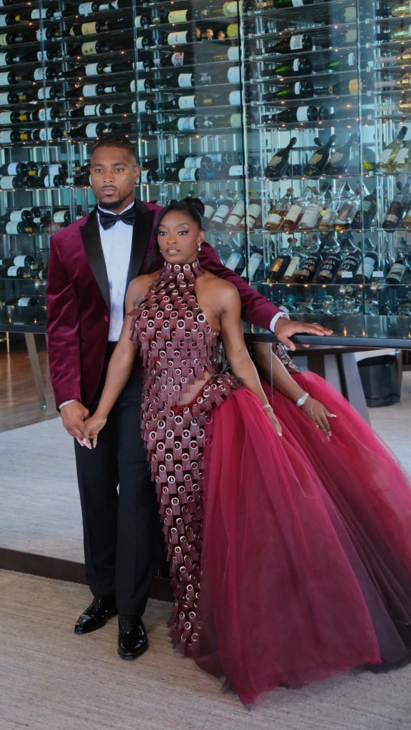 Simone Biles and husband Jonathan Owens pose in formal wear in front of a wine cellar display.