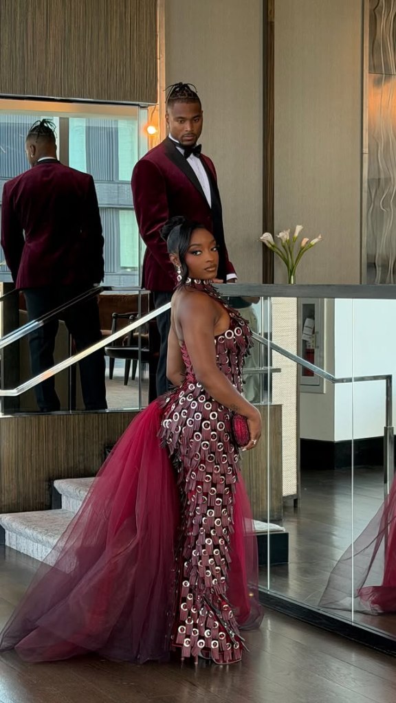 Simone Biles posing in a dark red gown with a man in a matching tuxedo behind her.
