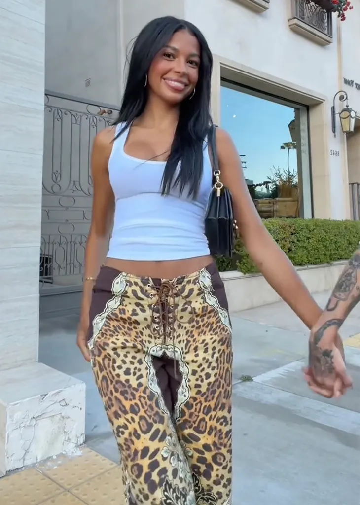 Layla Taylor in a white tank top and leopard print pants, holding hands with an unseen person, teasing a new girlfriend.