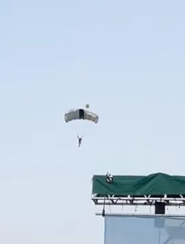 A person parachuting with an open parachute in the sky, with part of a building with a green roof visible in the bottom right corner.