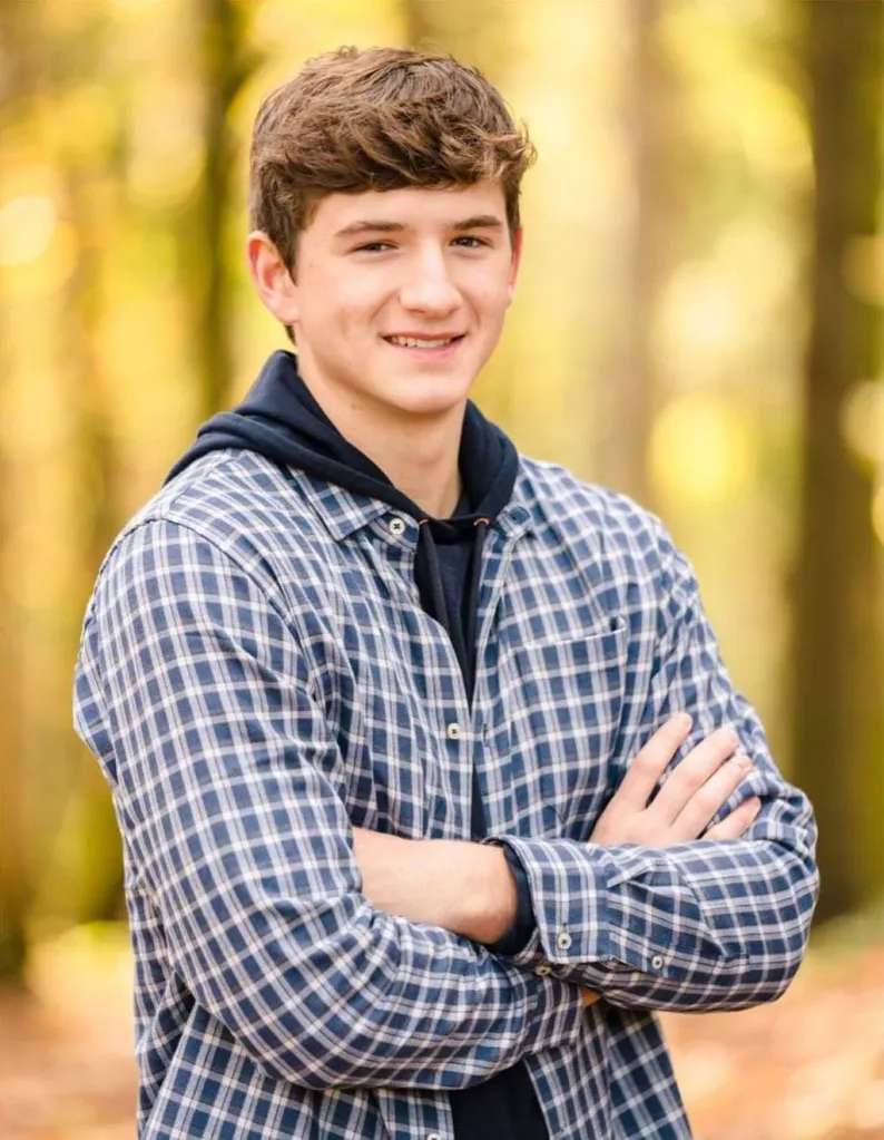 A young man with brown hair smiles at the camera, wearing a blue plaid shirt and hoodie with his arms crossed.