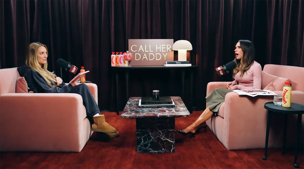 Victoria Beckham, in a pink top and grey skirt, being interviewed by Alexandra Cooper, in a black hooded top, black pants and brown boots, on the podcast