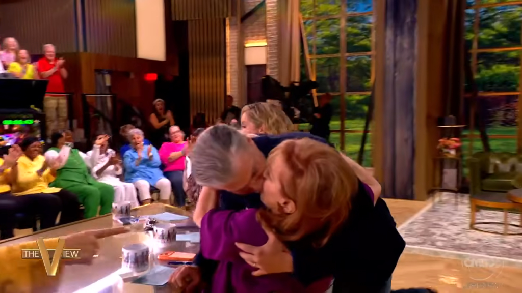 Anson Mount and Joey Behar share a kiss on live television, with an audience reacting in the background.