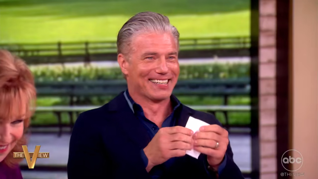 Anson Mount with gray hair smiles while holding a small piece of paper.