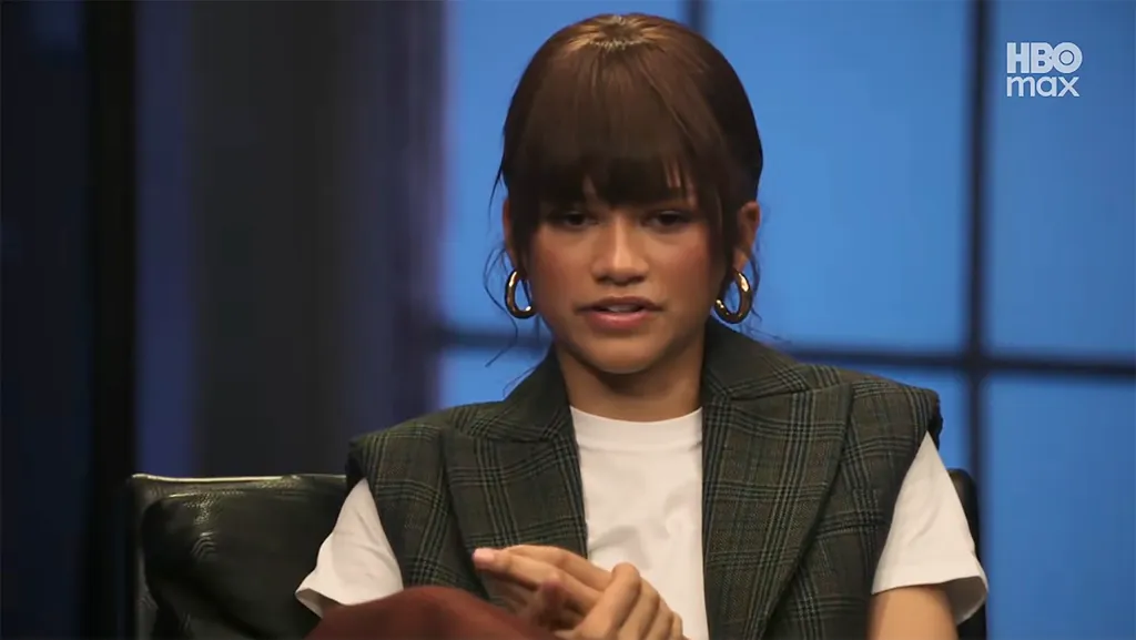 Zendaya in a dark plaid vest over a white t-shirt, large gold hoop earrings, and bangs, speaking during an HBO Max special.