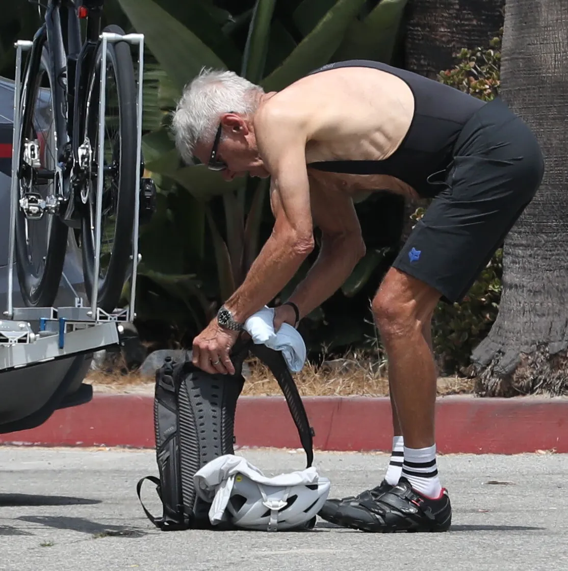 Harrison Ford in cycling attire bending over a backpack and helmet.