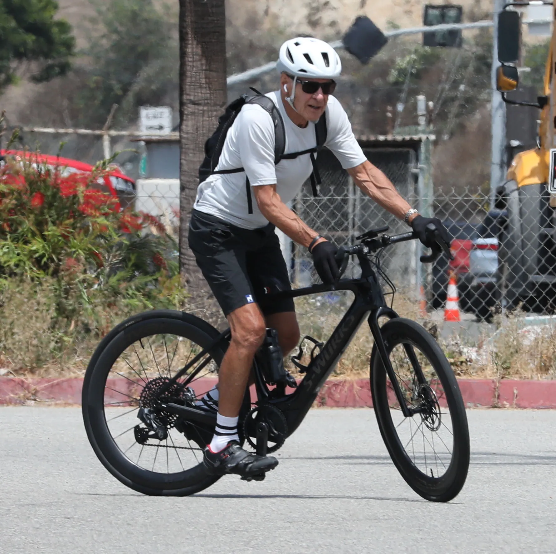 Harrison Ford, in a white long-sleeved shirt and black shorts, walking away from a car with bicycles on a bike rack.