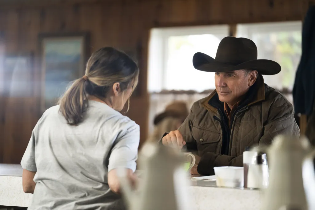 Kevin Costner as John Dutton in a quilted jacket and cowboy hat, talking to a woman from his seat at a table.