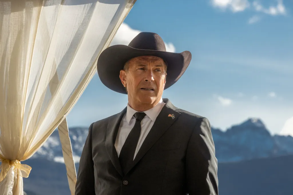 Kevin Costner in a black cowboy hat and suit, with a mountain range in the background.