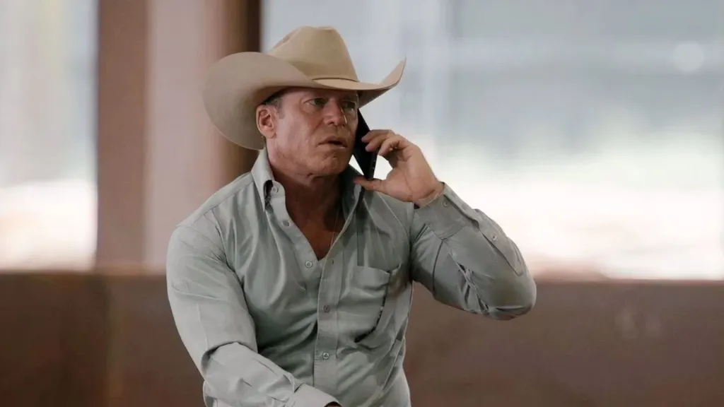 Taylor Sheridan as Travis Wheatley in Season 5 of Yellowstone, wearing a cowboy hat and light green shirt while talking on a phone.