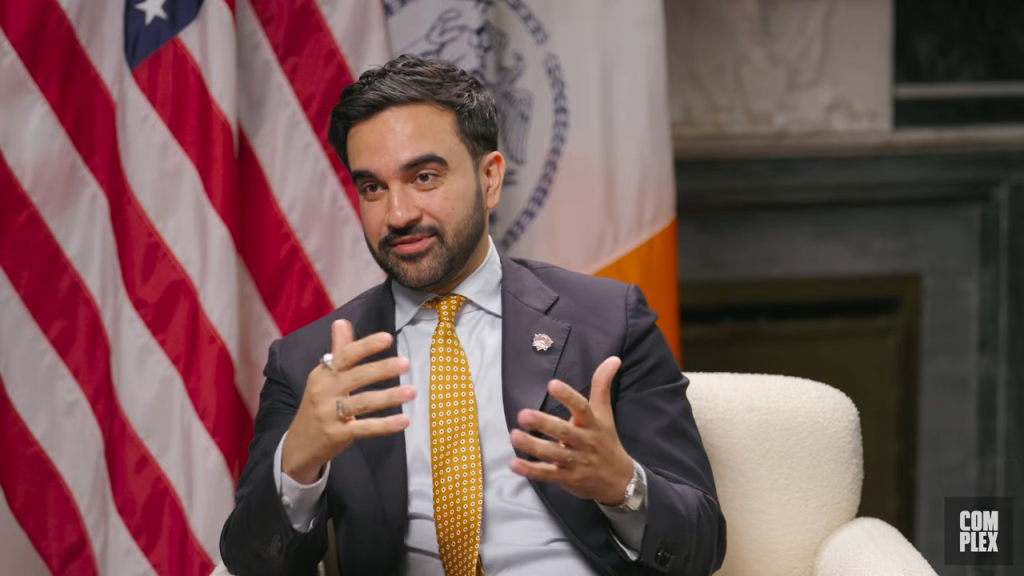 Zohran Mamdani speaking in front of an American flag and the New York City seal.