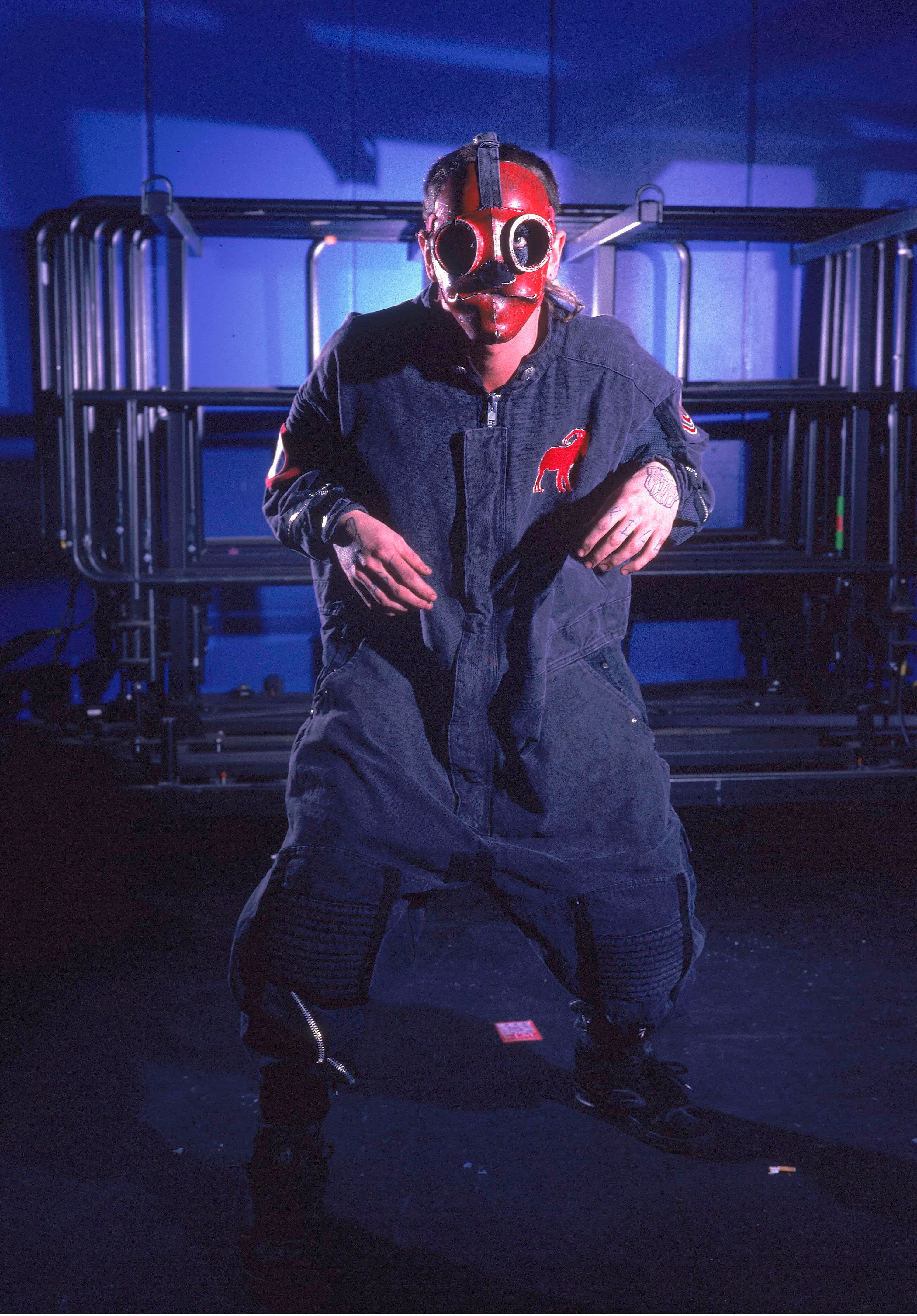 Sid Wilson in a red and black gas mask, dark jumpsuit with a red dog logo on the chest.