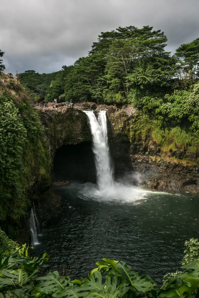 Rainbow Falls in Hilo, Hawaii, with lush green foliage and people watching from the top.