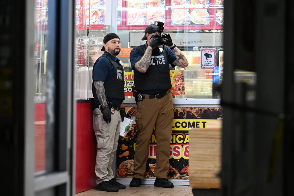 Two plainclothes NYPD officers, one with tattoos visible on his arms taking photos, are at the scene of a double shooting in Brooklyn.