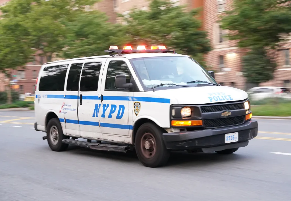 A white NYPD police van with blue stripes, emergency lights, and 