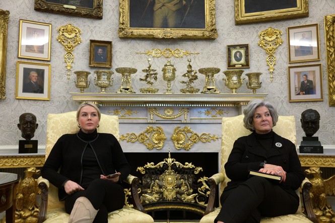 White House Press Secretary Karoline Leavitt and White House Chief of Staff Susie Wiles sit in chairs in front of a much-adorned fireplace and wall in the Oval Office.