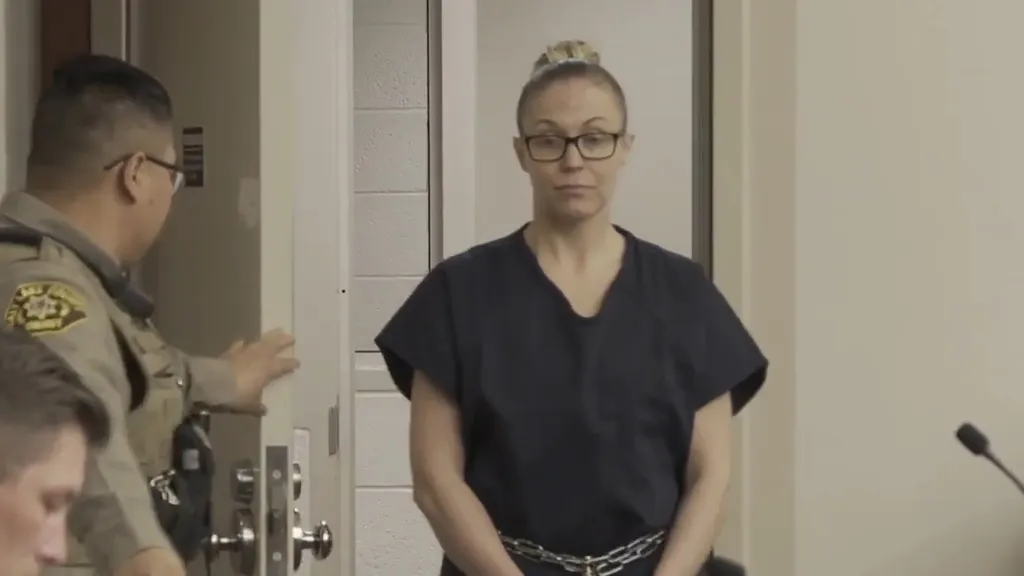 A woman in a navy shirt and glasses is led by a police officer into a room.