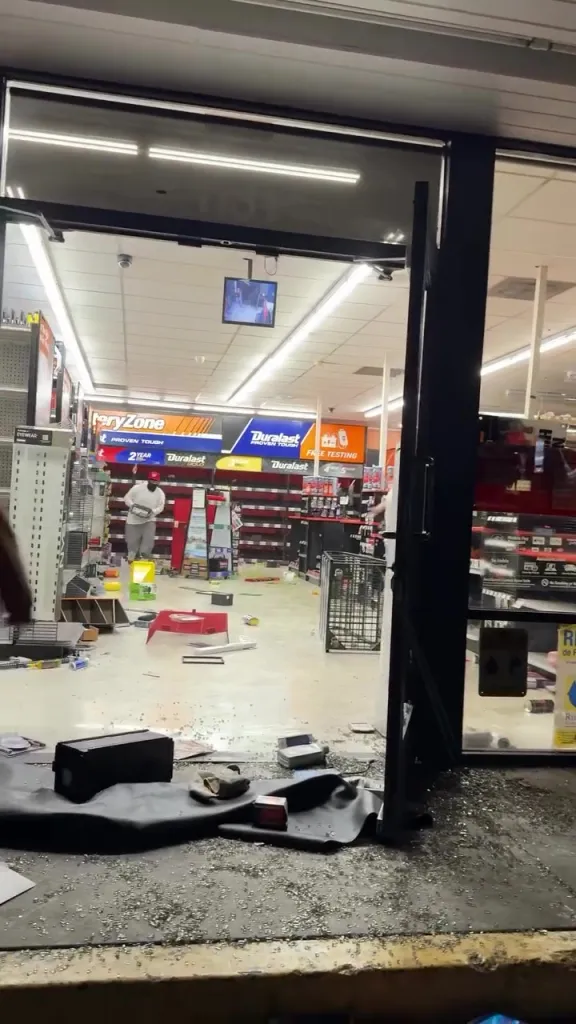 The interior of an AutoZone store appears ransacked, with empty shelves, debris on the floor, and a person walking towards the rear.