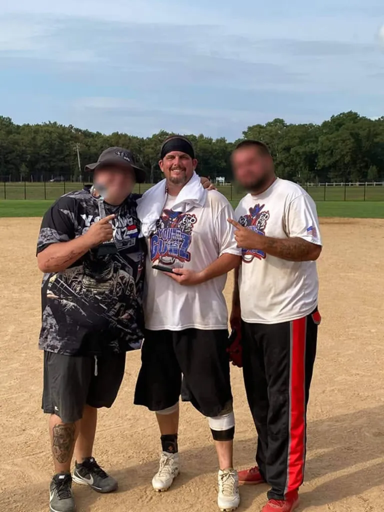 Nick Zavesky holding a trophy, flanked by two other men pointing at him, on a baseball field.