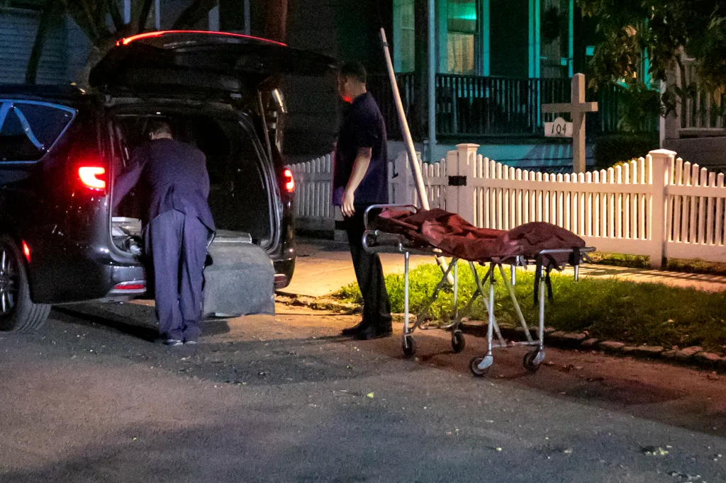 Two men standing next to a black minivan and a stretcher outside a house with a white picket fence at night.