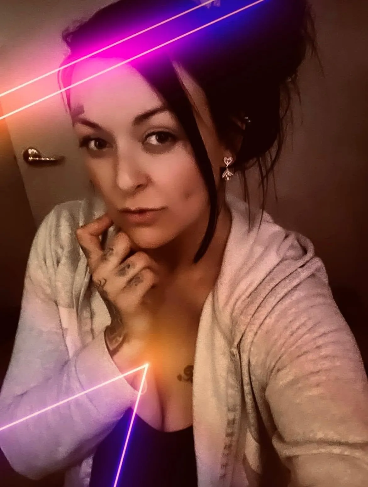 A woman with dark hair in a messy bun, wearing a white hoodie and a black top, poses for a selfie with one hand under her chin, surrounded by neon pink and purple light effects.