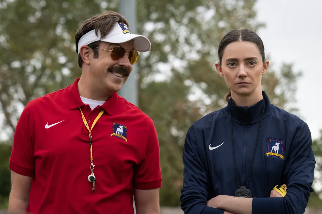 Ted Lasso smiling at a football field, while Coach Beard looks sternly at the viewer.