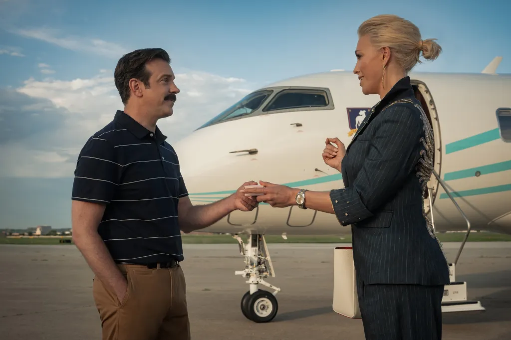 Ted Lasso gives a pink box to Rebecca Welton on an airport tarmac in front of a private jet.