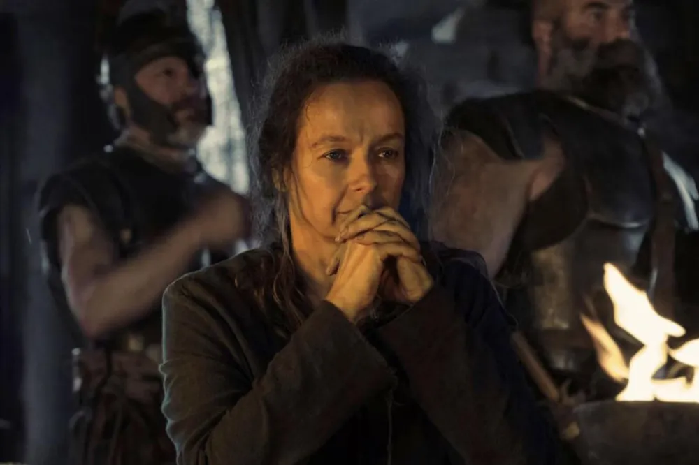 A woman with dark hair and a serious expression, hands clasped, in a dark setting with two men in armor behind her and a fire in the foreground.