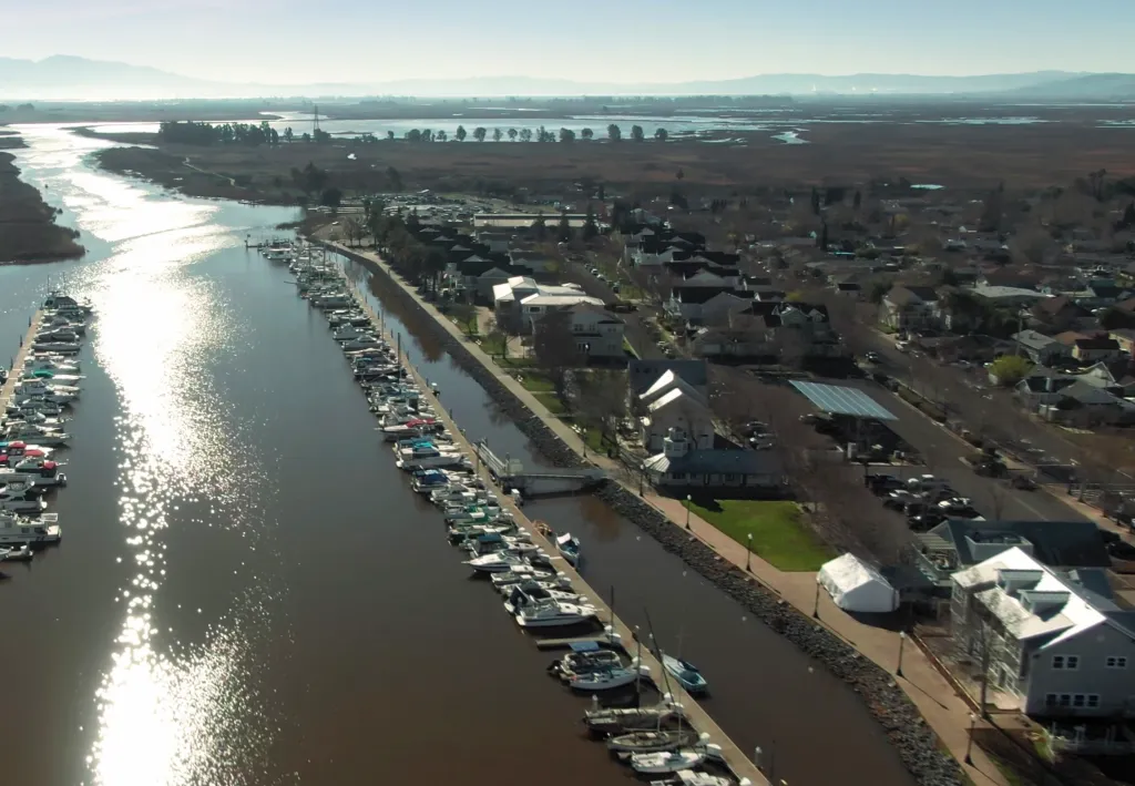 Aerial view of a marina with many boats docked on both sides of a river, with buildings and a town beyond.