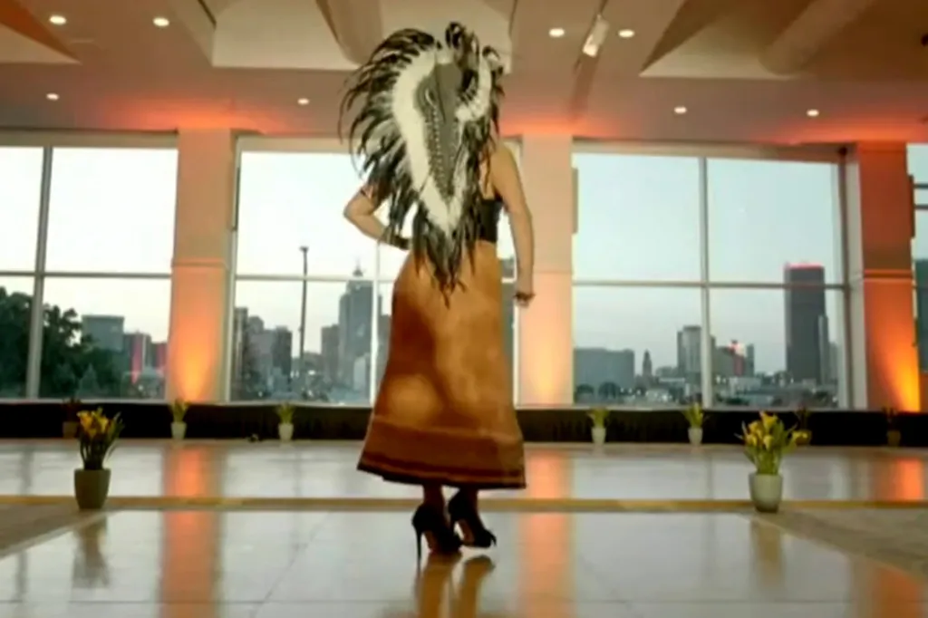 Woman in a feathered headdress and long skirt dancing in a room with large windows overlooking a city skyline.
