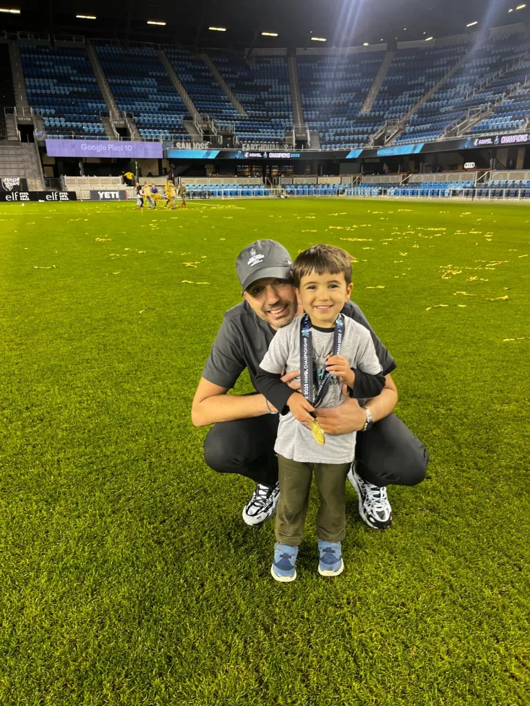 A man and a young boy holding a gold medal and smiling on a soccer field.