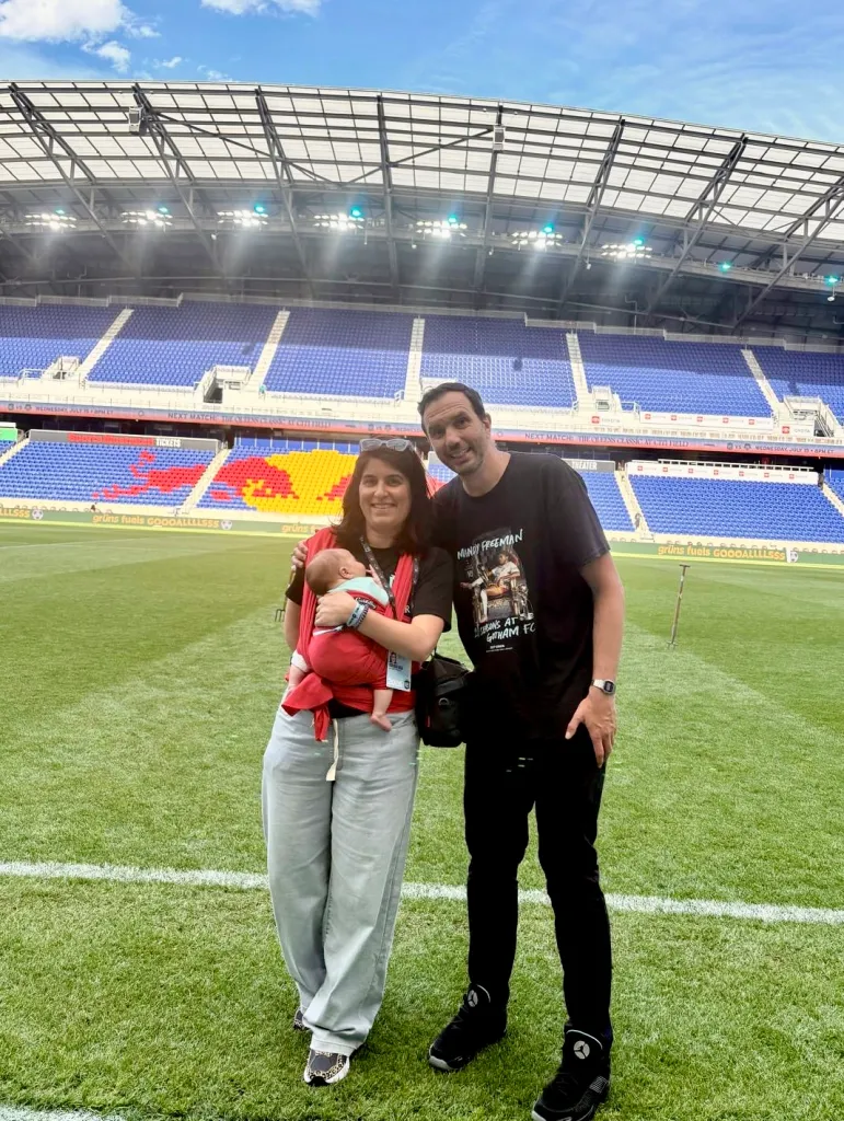 A family of three, including an infant, poses on a soccer field in front of stadium seating.