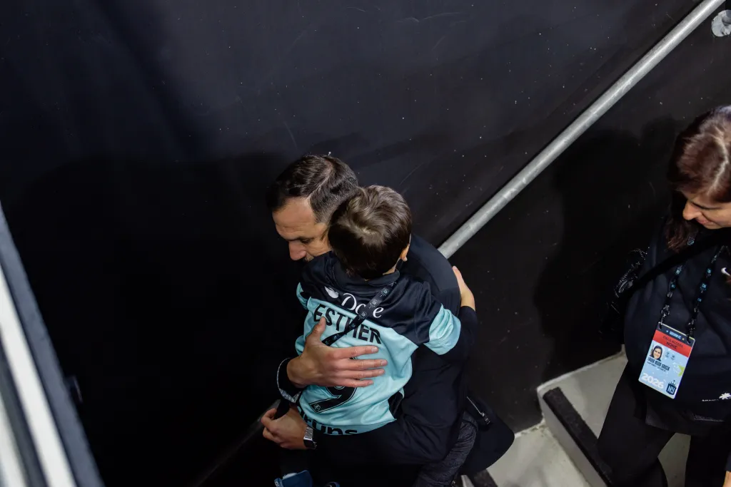 Man in black shirt holding a child in a turquoise and black jersey that says 